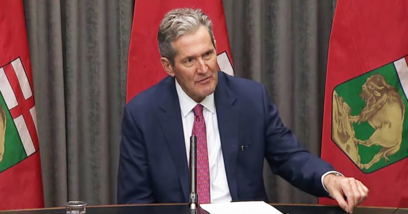 Premier Pallister at table with flags behind